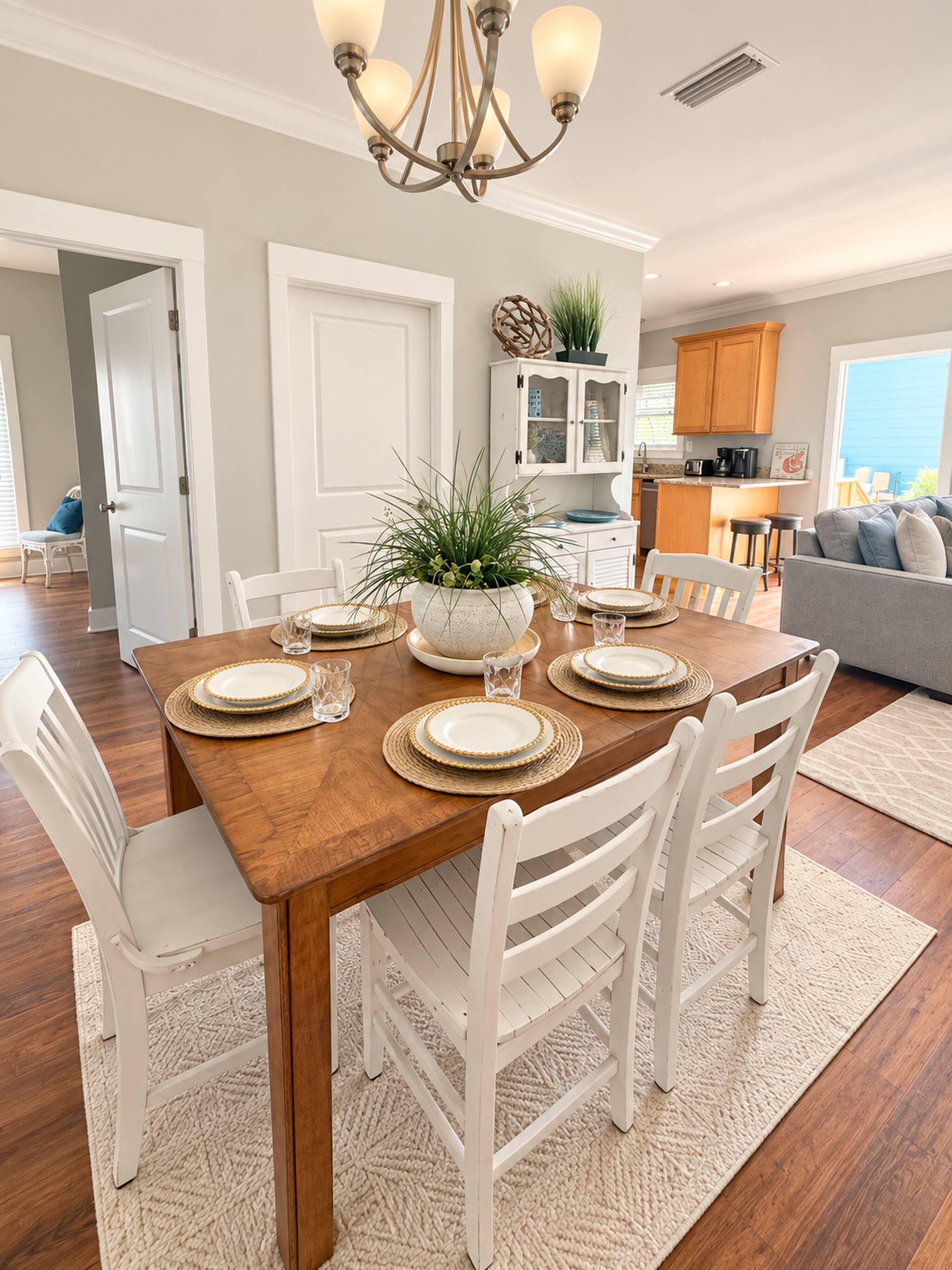 A dining area with a wooden table set for four, surrounded by white chairs, a chandelier, and a potted plant.