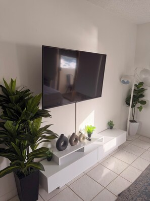 Un salon moderne avec un téléviseur à écran plat fixé sur une console blanche, entouré de plantes en pot et de vases décoratifs.