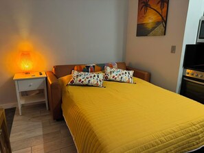 Une chambre à coucher comprenant un lit, une table de chevet, une lampe, un tableau et un coin cuisine.