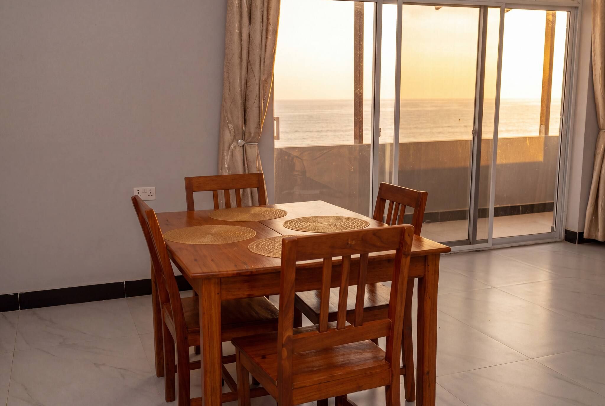 A dining area with a wooden table and chairs, a view of the sea through large glass doors, and a tiled floor.