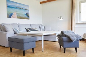 A living room with a sofa, ottomans, a coffee table, a lamp, and a painting of a dock. - Holiday home in Rimsting with swimming pool (Rimsting)