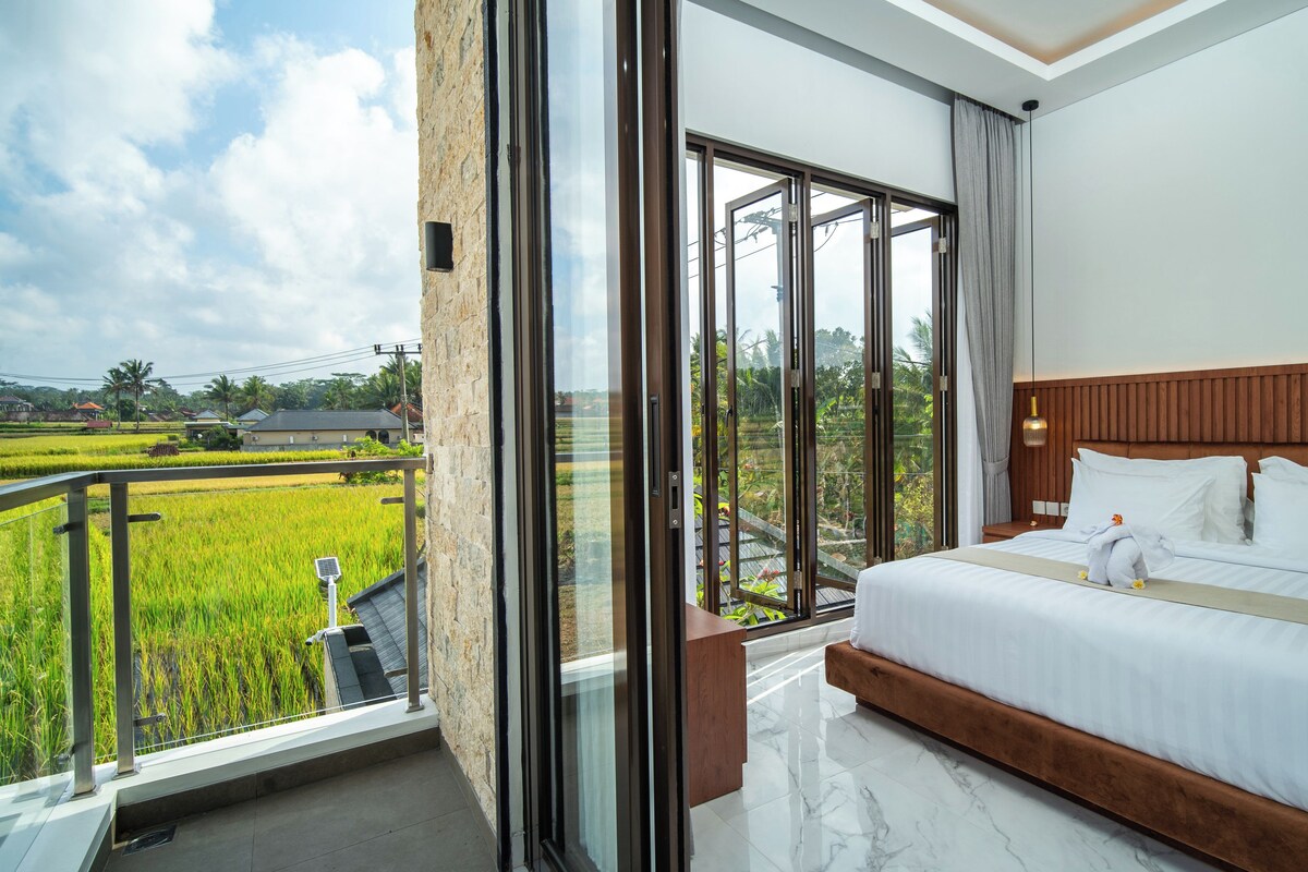 Une chambre d’hôtel avec un grand lit, une tête de lit en bois et un balcon donnant sur un paysage verdoyant.