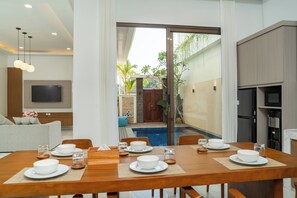 Un coin repas avec une table en bois dressée pour six personnes, un téléviseur fixé au mur et une vue sur une piscine et la verdure environnante.