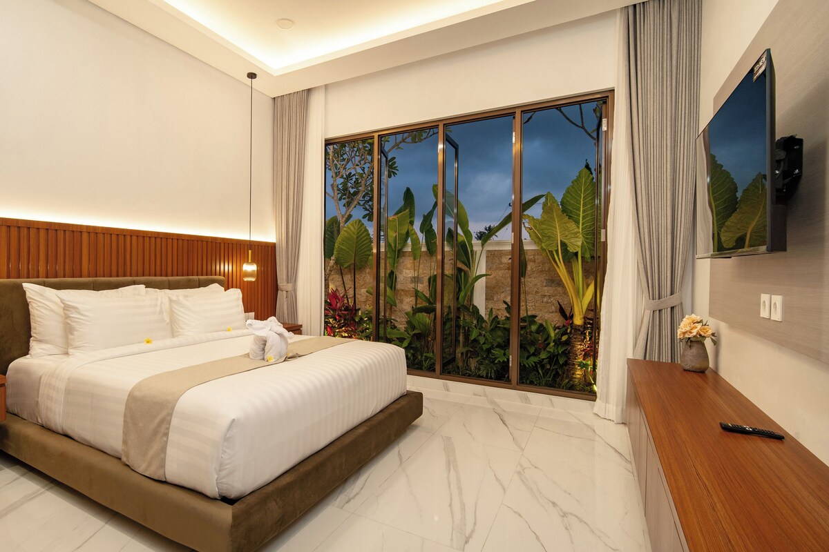 Une chambre d’hôtel moderne avec un grand lit, une tête de lit en bois, un téléviseur fixé au mur et une vue sur un jardin par la fenêtre.