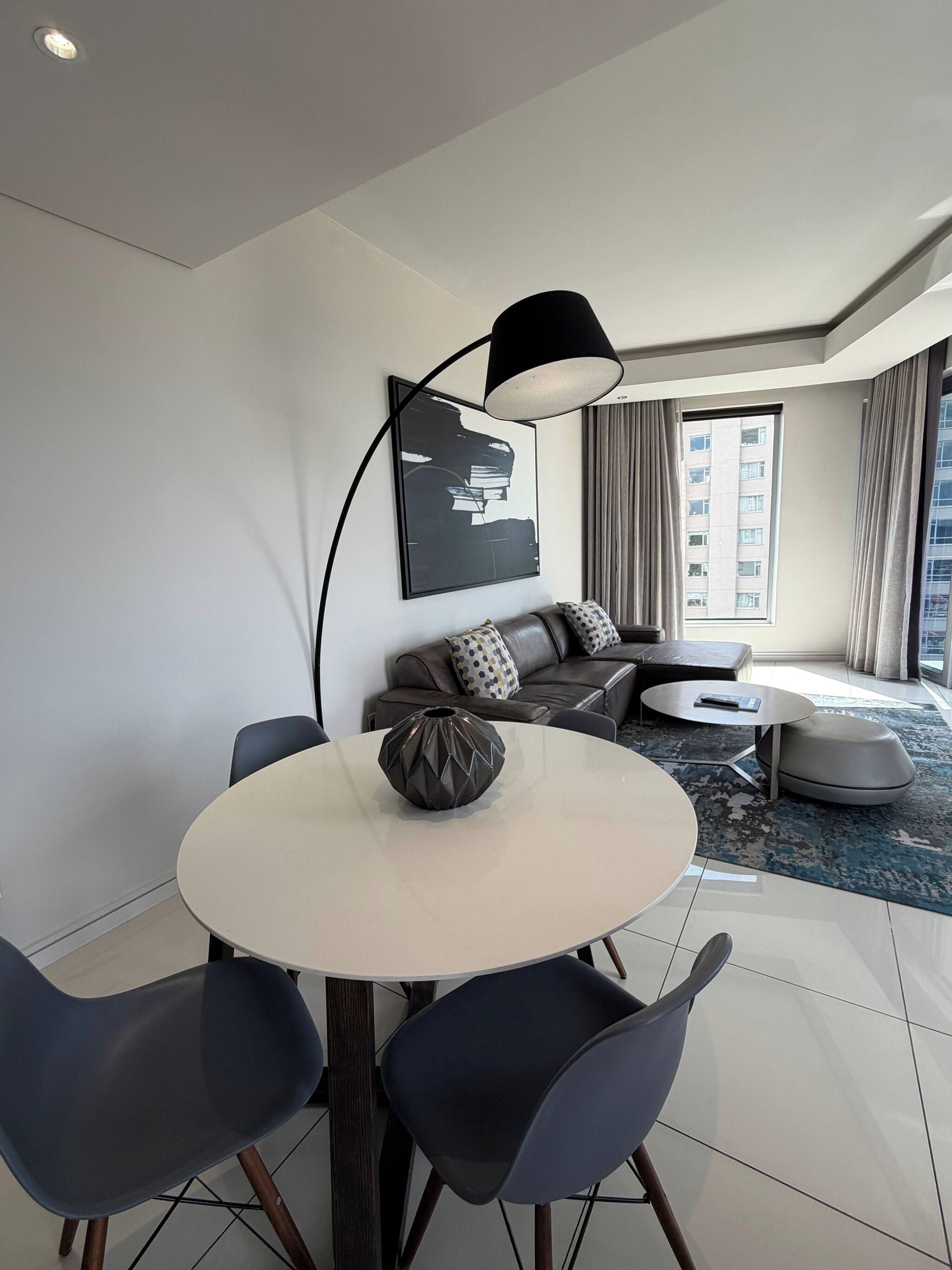 A modern living room with a round dining table, dark chairs, a sofa set, a large window with curtains, and a black floor lamp.
