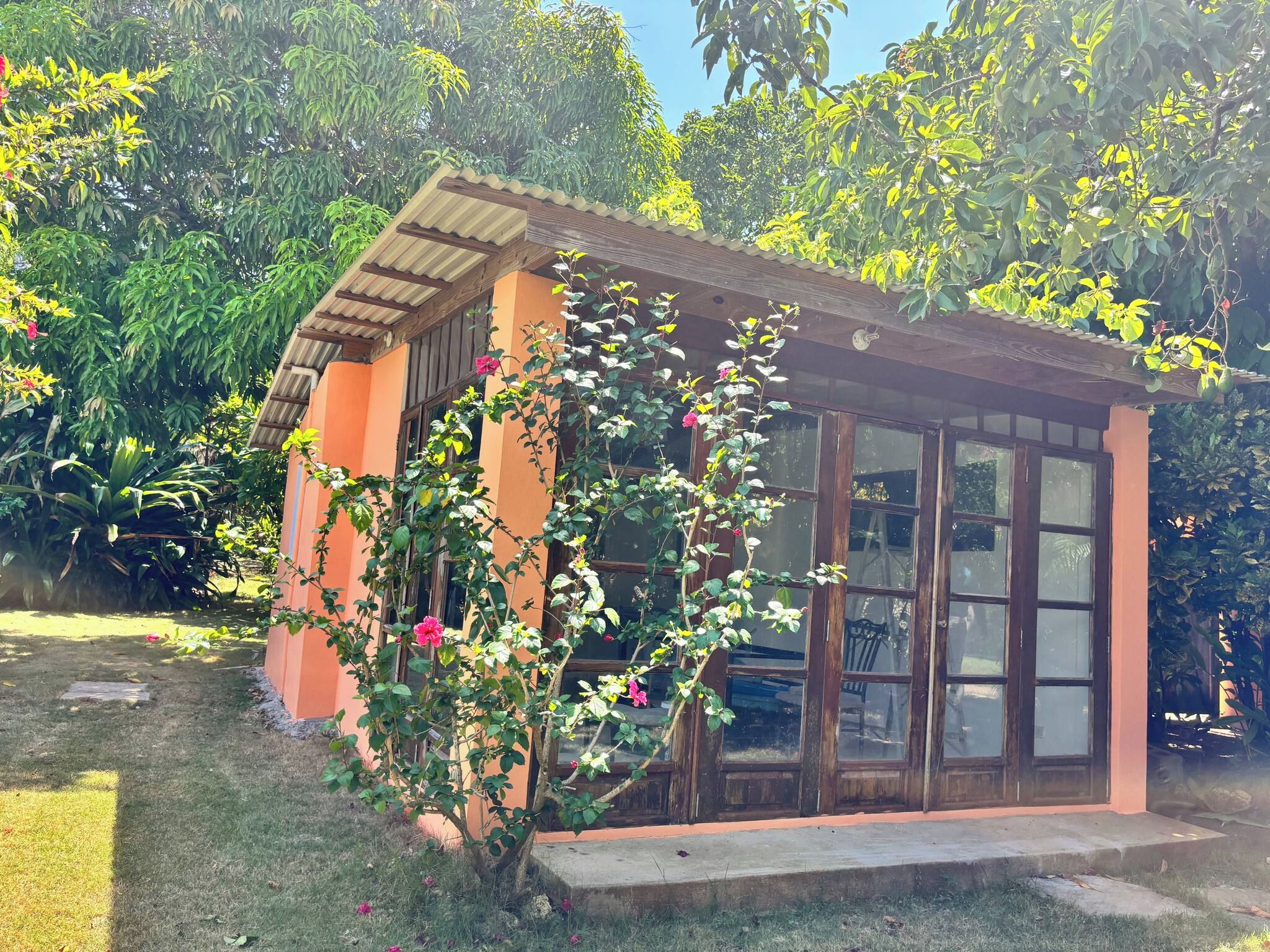 Un petit bâtiment d’un seul étage, doté d’un toit en pente, entouré d’une végétation luxuriante et de plantes en fleurs.