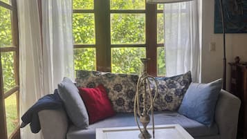 A room with a sofa, a coffee table, and a lamp. There are windows with curtains, and a painting on the wall.
