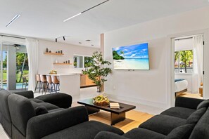 Una sala de estar moderna con un televisor de pantalla plana grande, un comedor con sillas y una cocina visible a través de una puerta abierta.