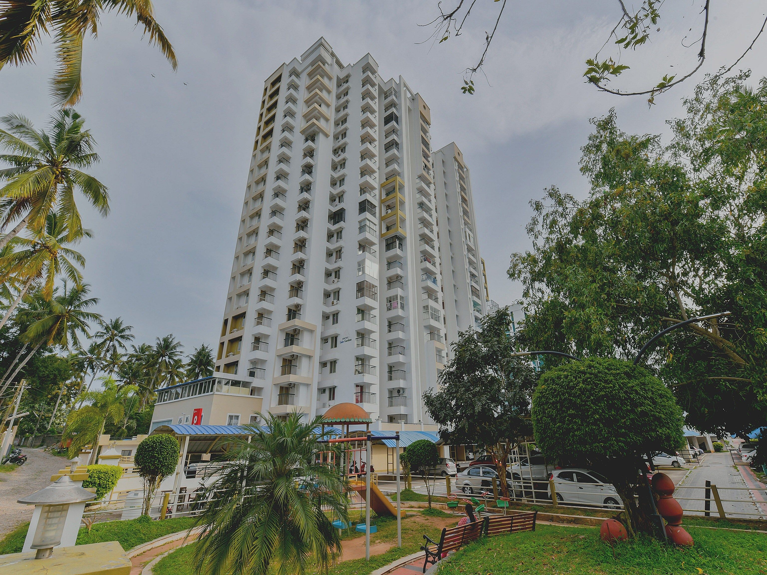 Edifício residencial alto, com várias varandas, cercado por áreas verdes e um parquinho.