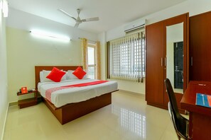Ein Hotelzimmer mit Bett, Schreibtisch, Stuhl, Kleiderschrank, Deckenventilator und einem Fenster mit Jalousien.