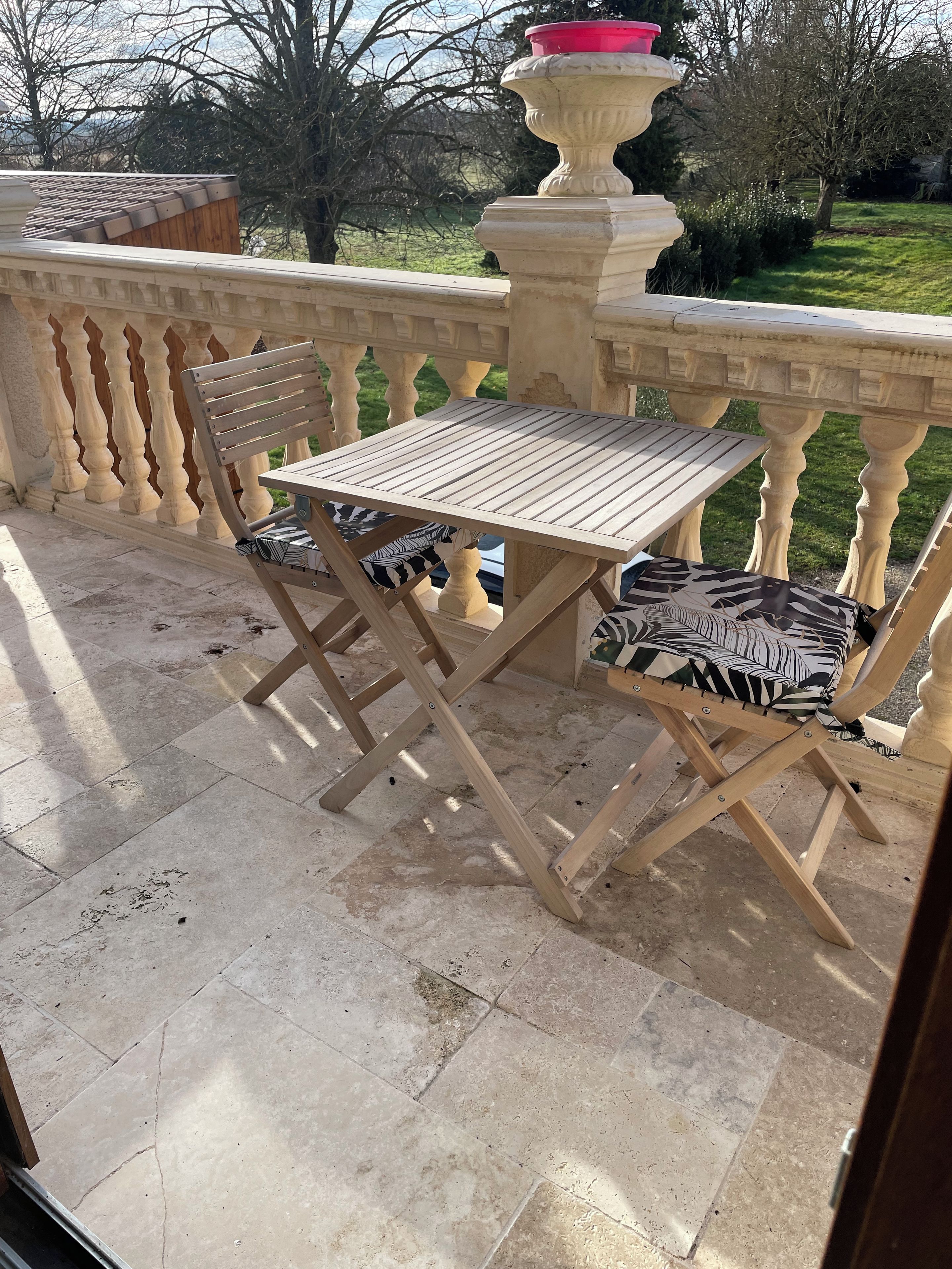 Un balcón con mesa y sillas de madera, una urna decorativa y vistas al jardín.