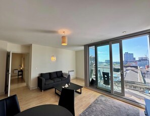 A modern living room with a black sofa, a coffee table, and a large window offering a city view. - Free Carpark Modern Lux 2 Bedroom Apt City Centre (Manchester)