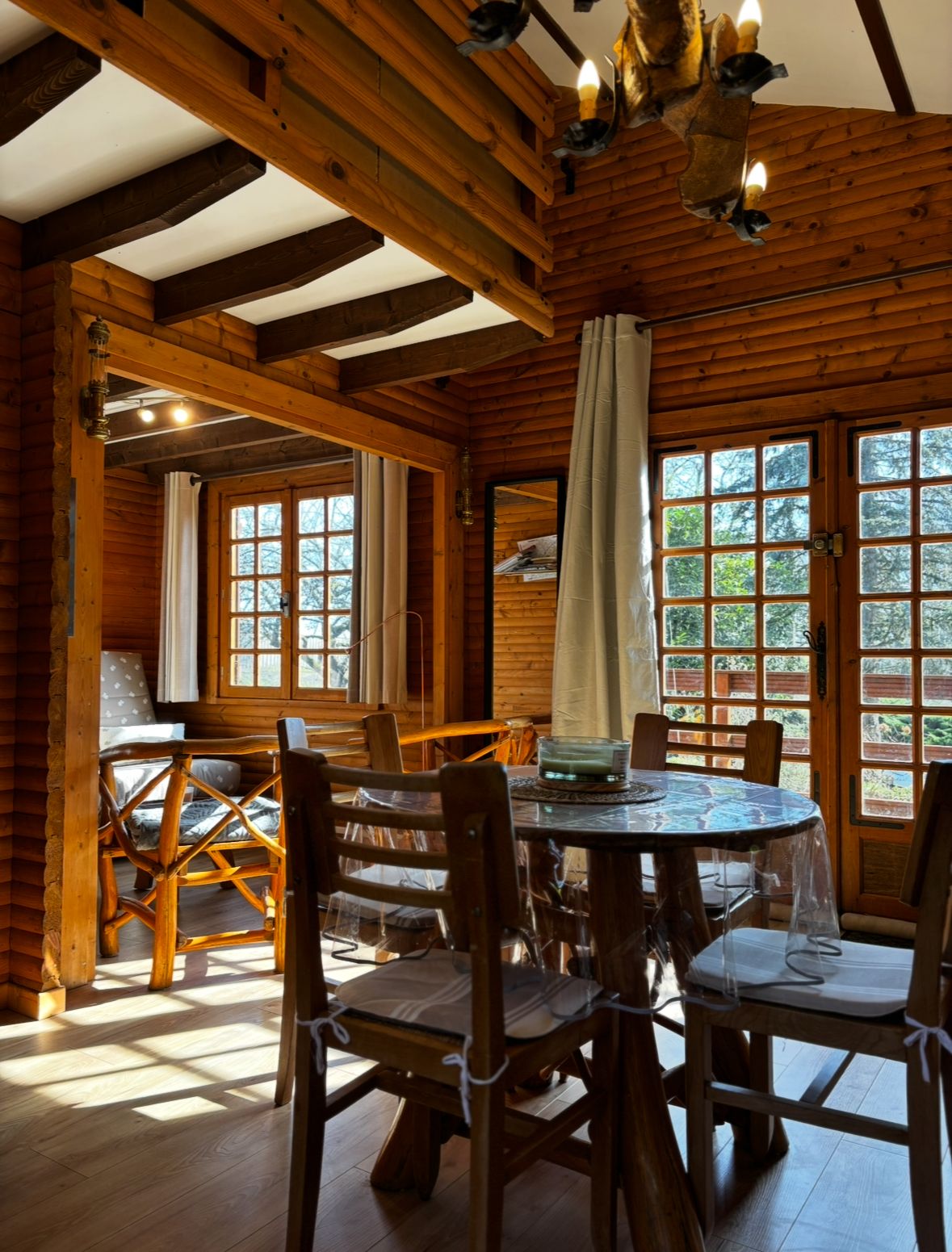Interior de una cabaña de madera con una mesa de comedor rodeada de sillas, ventanas amplias y un techo con vigas a la vista.