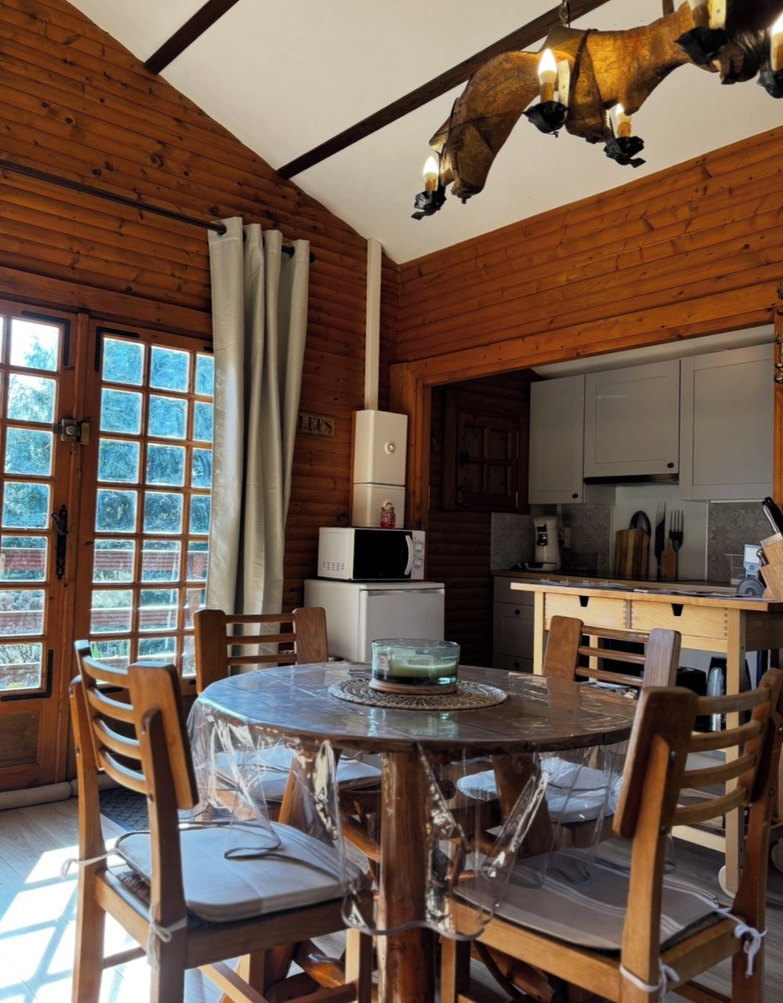 Interior em madeira com mesa de jantar redonda, cadeiras, área de cozinha e uma janela grande com cortinas.