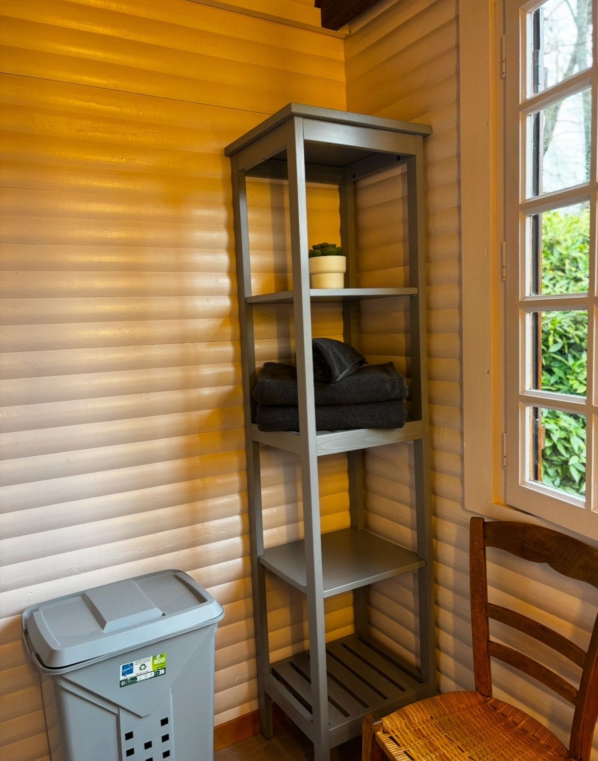 Una habitación de esquina con una estantería metálica gris, una silla de madera y un cesto de basura.