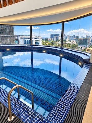 A modern indoor swimming pool with a curved design, large windows offering city views, and a metal ladder for access. - Capital M Residences - LEED Certified (Nairobi)