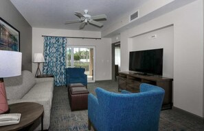 Ein Hotelzimmer mit einer Couch, zwei blauen Sesseln, einem Fernseher, einem Deckenventilator und Blick durch eine Glastür.