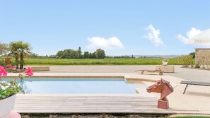 Zwembad met houten terras, een paardenbeeld en uitzicht op een groen veld en bomen.