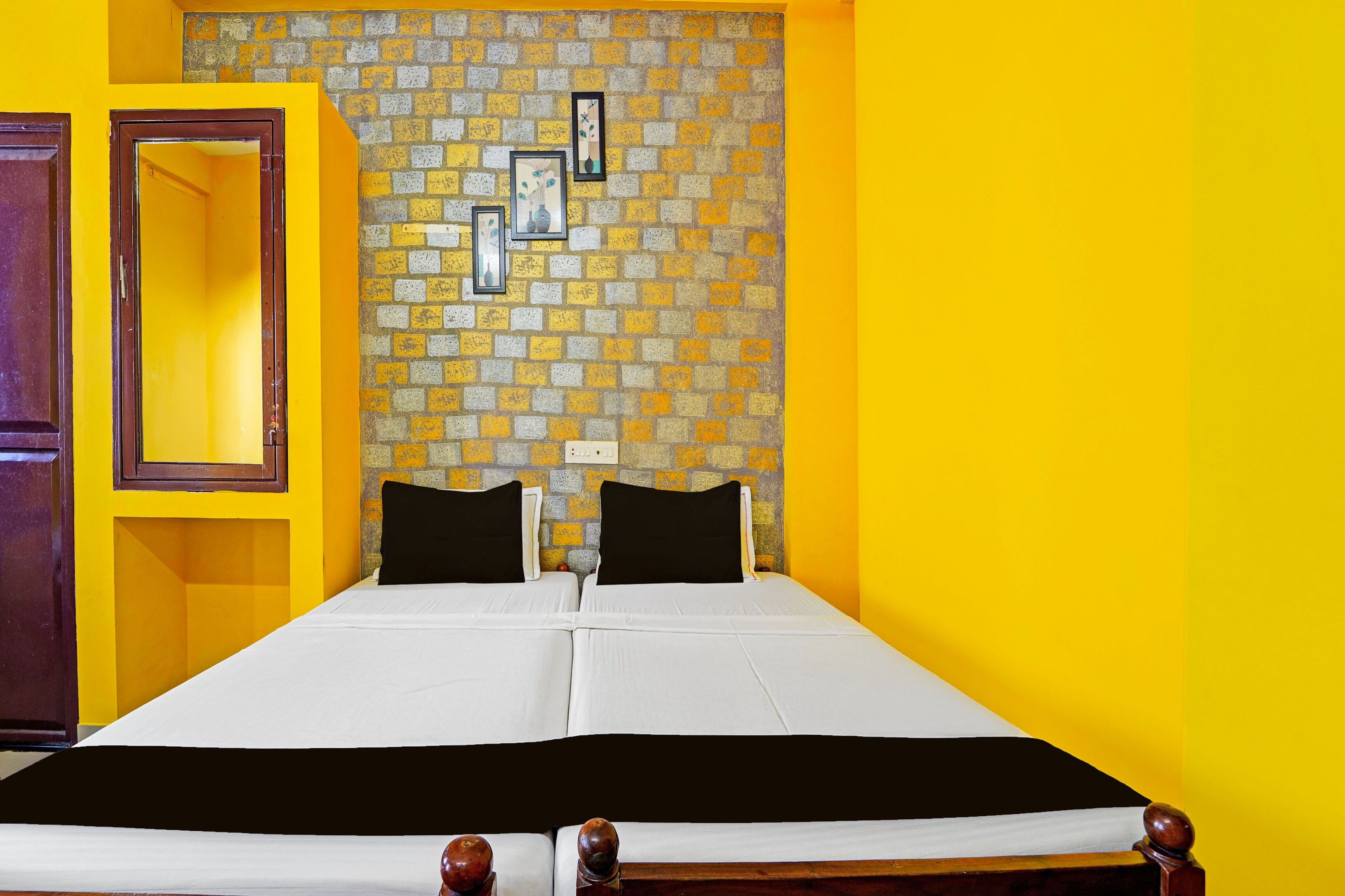 Cama de casal com roupa de cama branca e almofadas pretas num quarto com paredes amarelas e uma parede de destaque com padrão de tijolos.