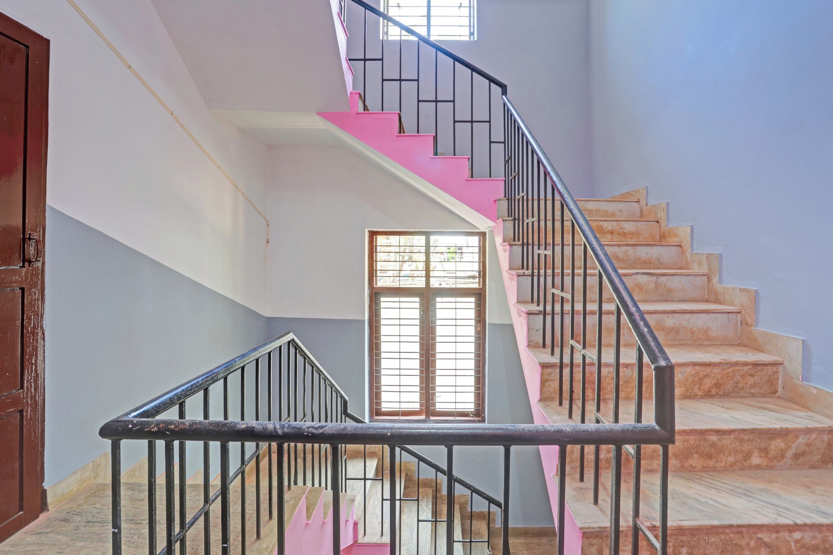 Uma escadaria com degraus cor-de-rosa e guarda-corpos pretos, que leva a um patamar com uma janela.