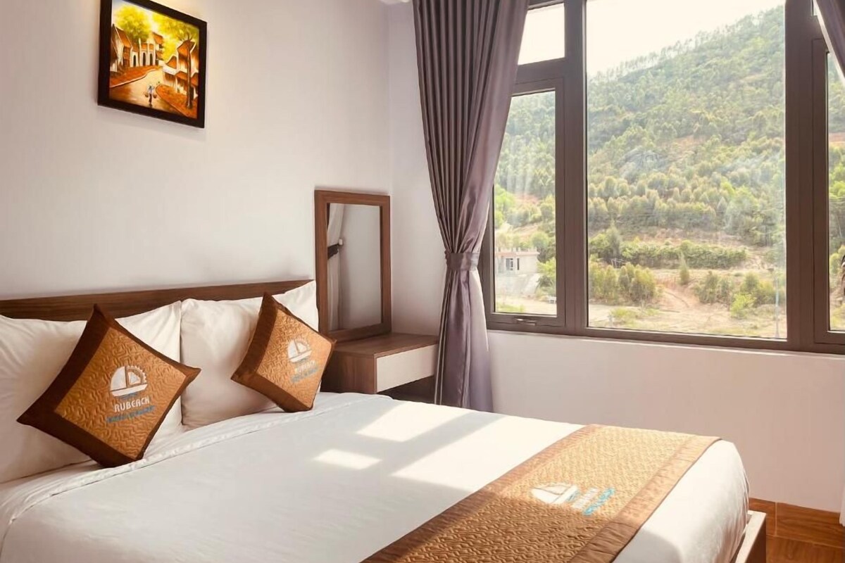 Une chambre d’hôtel avec un lit, un tableau au mur, une fenêtre avec des rideaux et une vue sur une zone boisée.