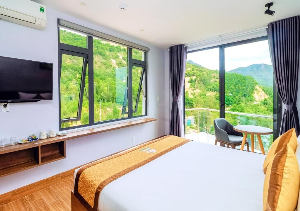 Habitación de hotel con una cama grande, televisor montado en la pared, ventana con vistas a zonas verdes y balcón con silla y mesa.
