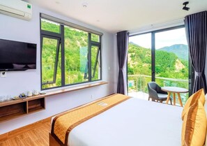 Habitación de hotel con una cama grande, televisor montado en la pared, ventana con vistas a zonas verdes y balcón con silla y mesa.