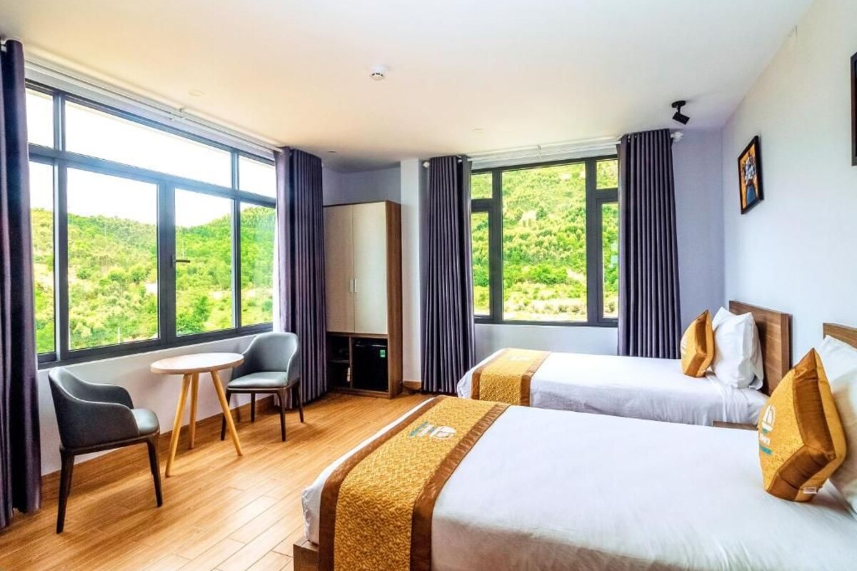 Une chambre d’hôtel avec deux lits, une petite table, une chaise et de grandes fenêtres donnant sur un paysage verdoyant.
