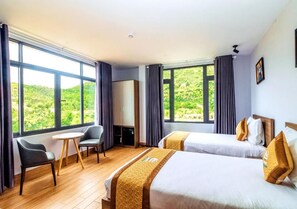 Une chambre d’hôtel avec deux lits, une petite table, une chaise et de grandes fenêtres donnant sur un paysage verdoyant.