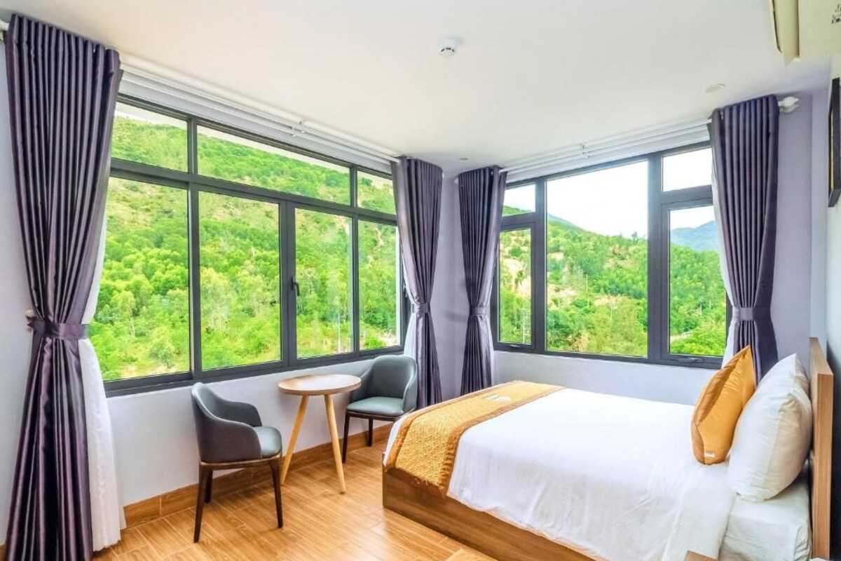 Une chambre d’hôtel avec un grand lit, une petite table, une chaise et une vue sur des collines verdoyantes, visible à travers de grandes fenêtres.