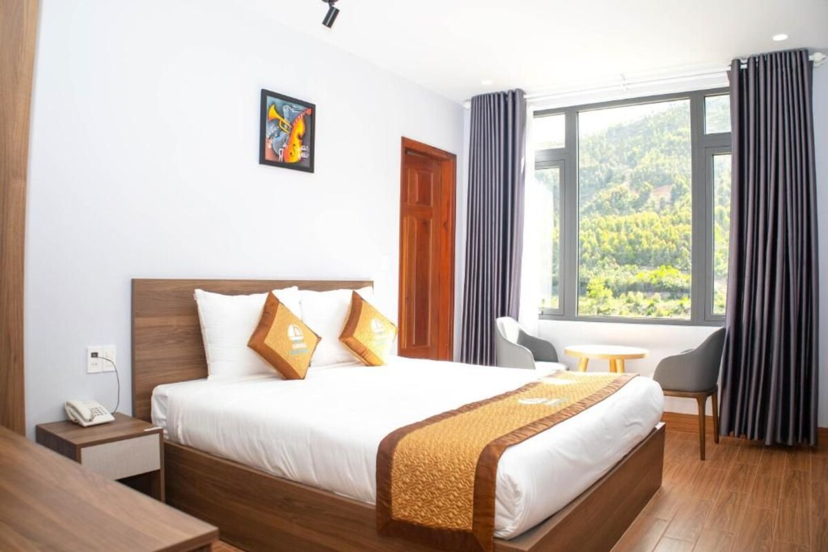 Une chambre d’hôtel avec un grand lit, une table de chevet avec un téléphone, une chaise, une petite table et une fenêtre avec des rideaux.
