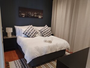 A neatly made bed with white bedding and patterned pillows, a nightstand with a lamp, a cityscape artwork on the wall, and a checkered rug on the floor. - Durbanville Work Home (Cape Town)