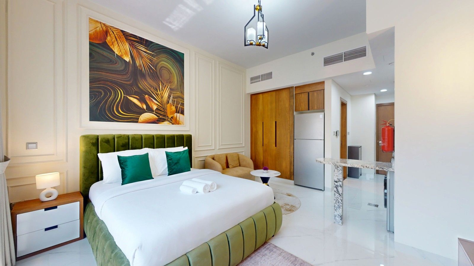 A modern hotel room with a large bed, a small round table, a kitchenette, and a painting on the wall.
