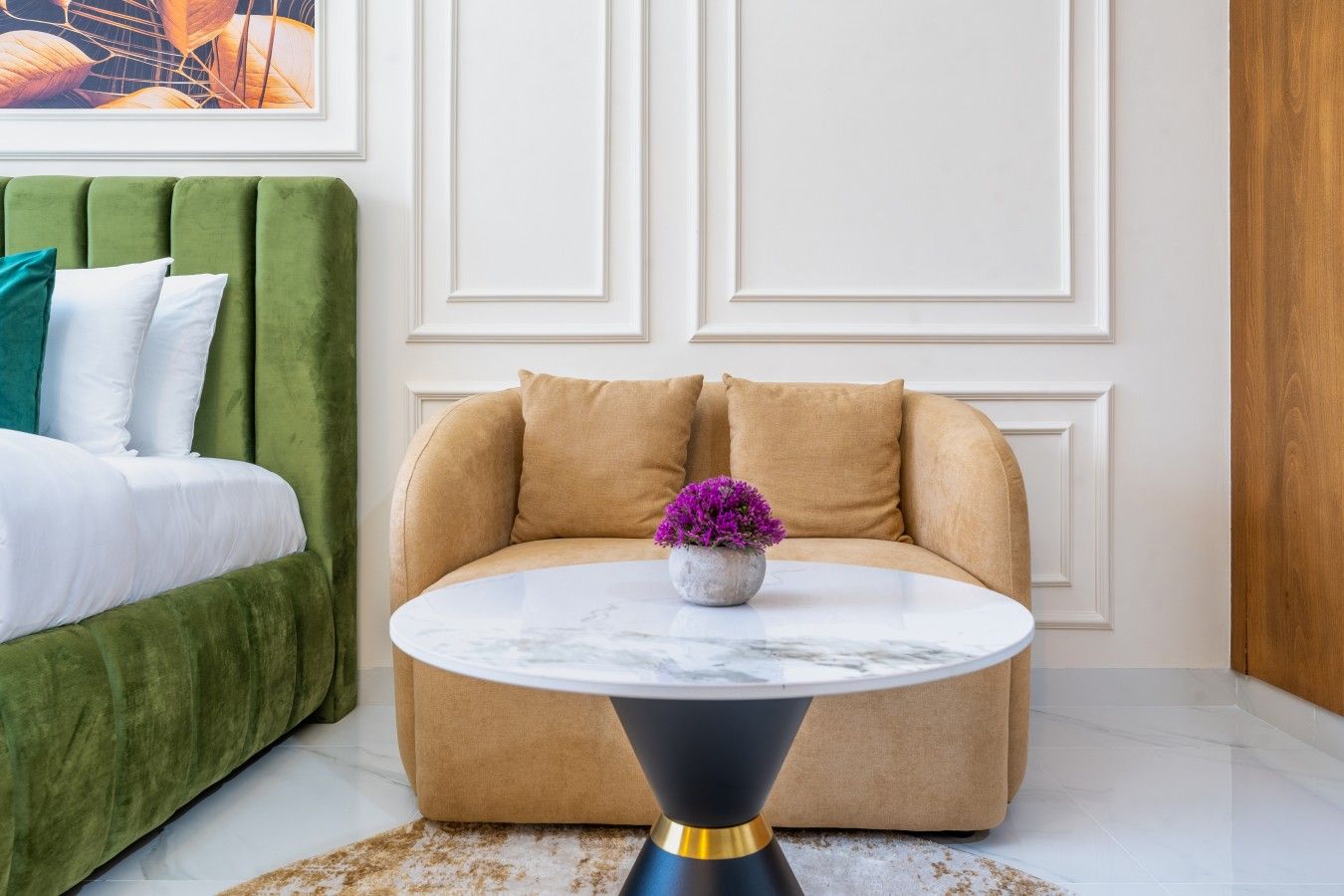 A modern living room with a green sofa, a round coffee table with a vase of purple flowers, and a white wall with paneling.