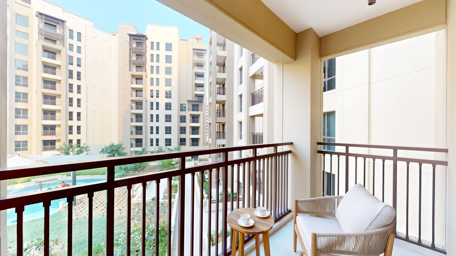 Un balcon donnant sur des immeubles de grande hauteur, une petite table avec une tasse et une soucoupe, et un fauteuil en osier.