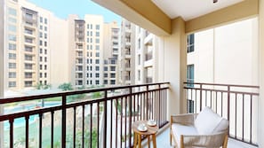 Un balcon donnant sur des immeubles de grande hauteur, une petite table avec une tasse et une soucoupe, et un fauteuil en osier.