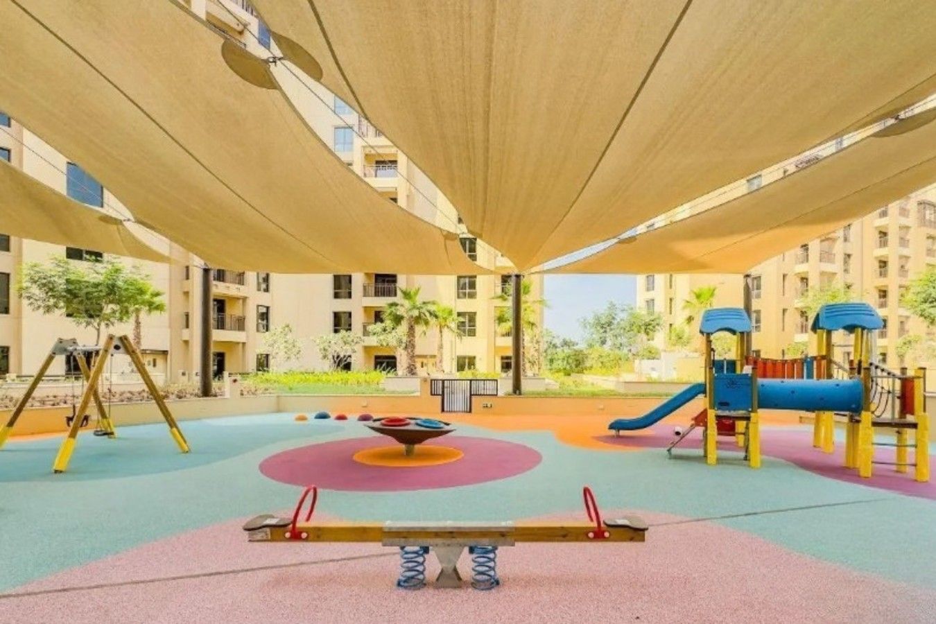 A playground with swings, a seesaw, and a slide, surrounded by apartment buildings.