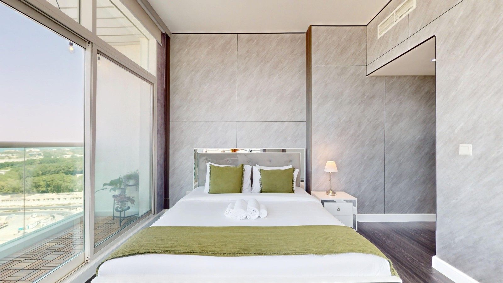 A modern hotel room with a large bed, a nightstand, a lamp, and a view of the city through a large window.