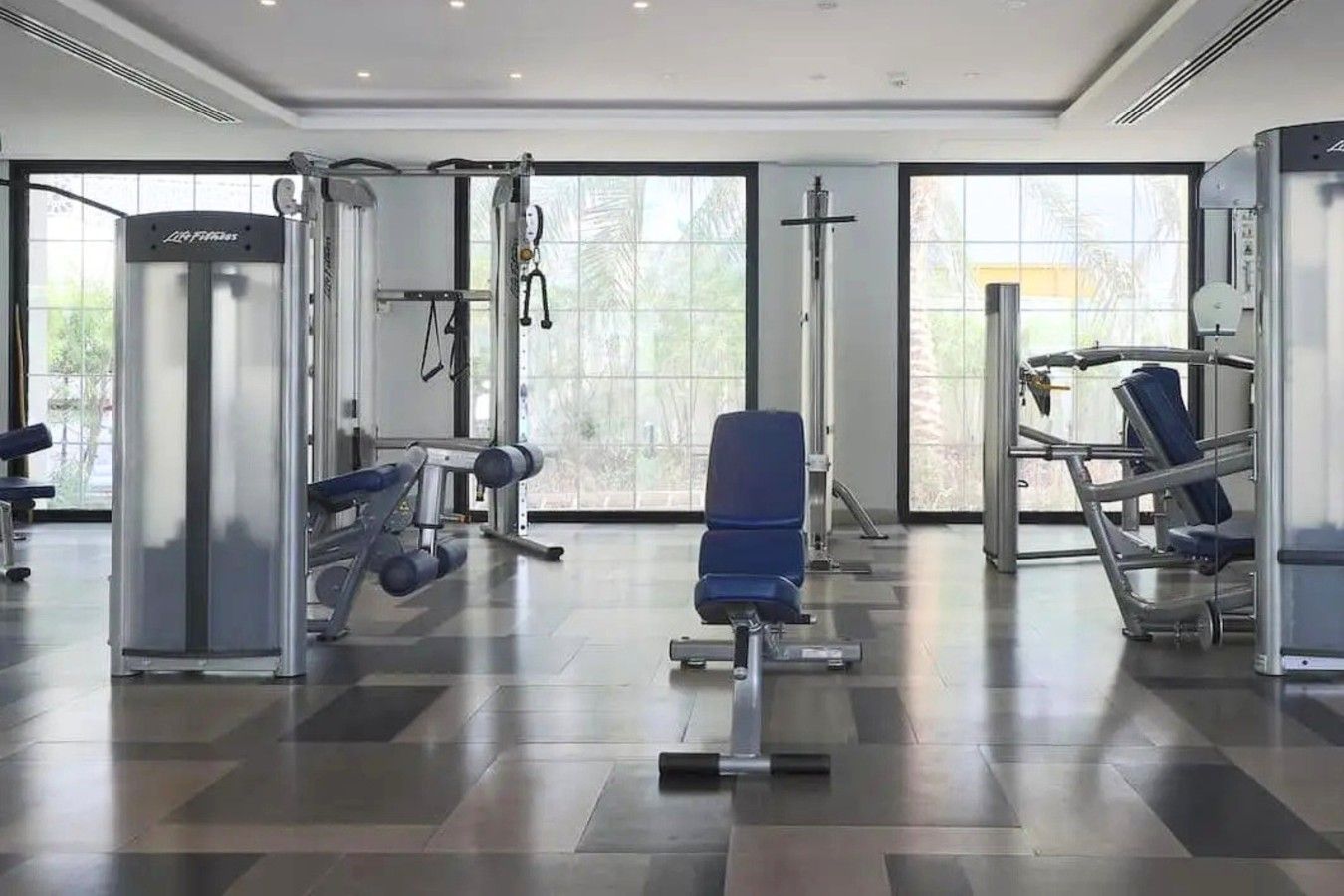Une salle de sport équipée de divers appareils d’exercice, dotée de grandes fenêtres et d’un sol carrelé.