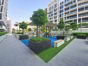 Un espacio exterior moderno con un parque infantil, zonas de estar y jardineras.