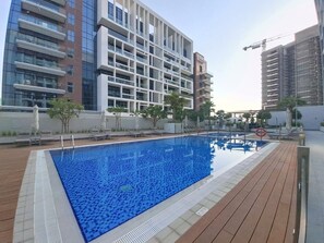Una piscina rectangular con un bordeado de baldosas, rodeada de edificios modernos y una grúa de construcción al fondo.