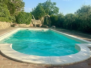 A rectangular swimming pool with a curved edge, surrounded by a stone wall and lush greenery. - New Listing (Camucia)