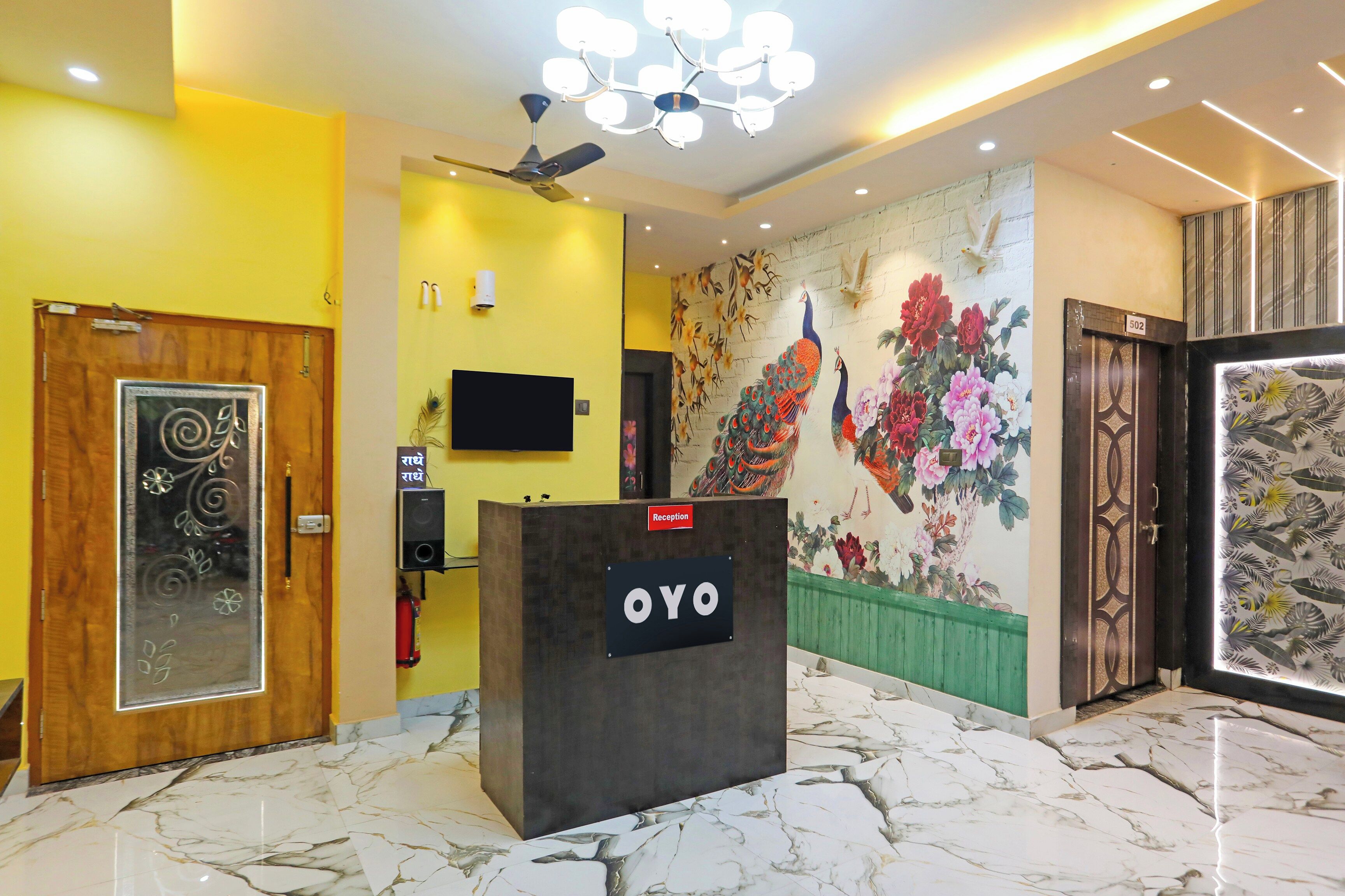 Un mostrador de recepción con un cartel, un ventilador de techo, un televisor montado en la pared y un mural con pavos reales y flores.
