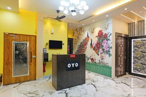 A reception desk with a sign, a ceiling fan, a wall-mounted TV, and a mural with peacocks and flowers. - Hotel O by OYO The Baba Guest House (Varanasi)