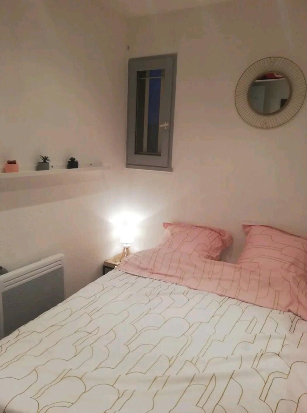 Une chambre à coucher avec un lit, une table de chevet, un miroir fixé au mur et une fenêtre avec un store fermé.