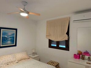 Une chambre à coucher comprenant un lit, une table de chevet, un ventilateur de plafond, une fenêtre avec un rideau, un climatiseur fixé au mur et un tableau encadré.