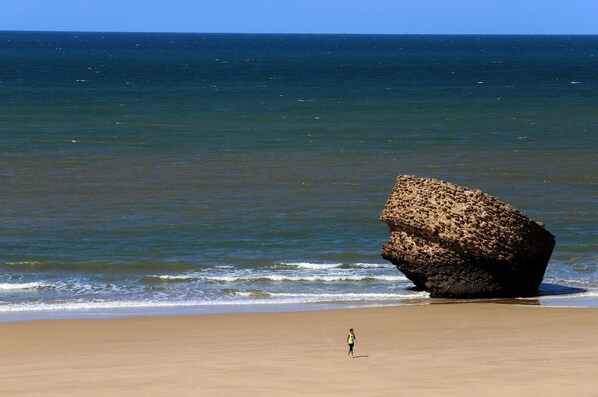 Une plage avec une grande formation rocheuse dans l’eau et une personne debout sur le sable.