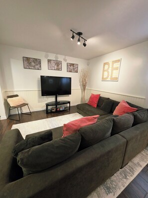 Ein modernes Wohnzimmer mit einem großen Flachbildfernseher, einem Ecksofa mit roten und schwarzen Kissen, einem weißen Teppich und einer Wandleuchte.