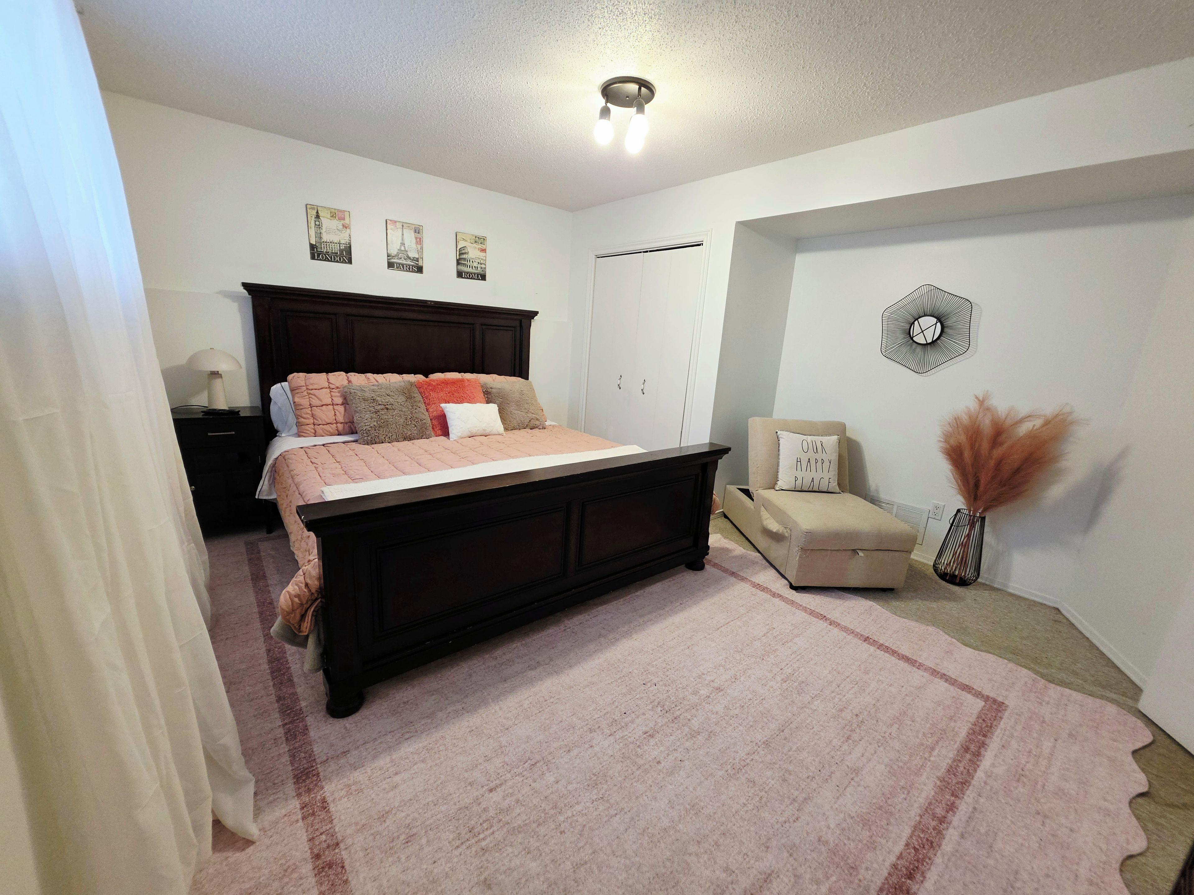 A bedroom with a dark wooden bed, a white nightstand, a beige armchair, a decorative wall piece, and a vase with pampas grass.