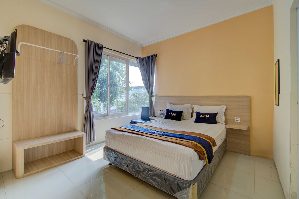A hotel room with a bed, a wooden wardrobe, a window with curtains, and a view of greenery outside.